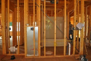 Interior stud walls framed around a molded tub and shower unit, wiring and pipe roughed in, with a worker turned away at the far side.