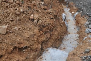 A drainage trench dug along the foundation wall and lined with stone and fabric, excavated clay heaped to one side below a garage door.