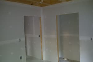 A drywalled room corner with taped and mudded seams, two door openings, and exposed floor framing overhead.