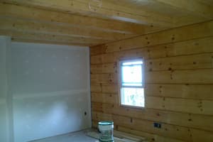An interior room where a finished log wall meets a taped drywall partition, a small window between beams and buckets on the bare floor.