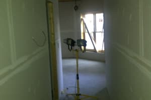 A hallway with drywall hung and seams taped, opening into a room lit by twin work lights on a yellow tripod stand.