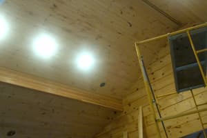 Looking up at a board ceiling with recessed lights burning above stacked log walls, scaffolding set against the wall below.