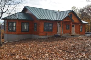 The front of a finished log home with a standing-seam metal roof and a covered porch, bare graded dirt and fallen leaves across the yard.
