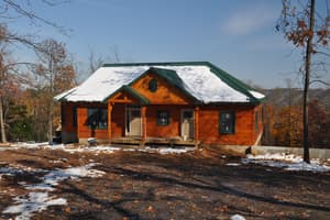 A finished log home on a ridge with snow lying on its metal roof, a plank porch deck out front and patchy snow on the graded yard.