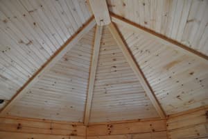 Looking up at the boarded underside of a hip roof, timber rafters meeting at a center post above the top of a log wall.