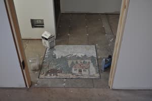A mosaic set into the tile floor — a mountain scene with a house among pines, laid in small stones.
