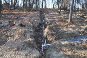 A narrow utility trench cut across cleared ground, white pipe running its length with branch fittings standing up and gravel to one side.