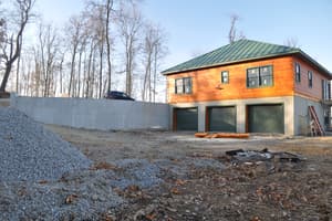 A finished walkout level with three garage doors below log siding and a metal roof, a concrete retaining wall and gravel piles in front.