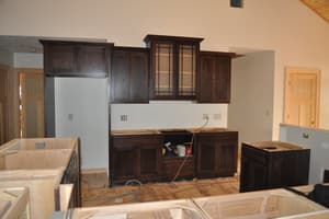 A kitchen with dark-stained upper and base cabinets hung but no countertops, unfinished island boxes and cords across the floor.