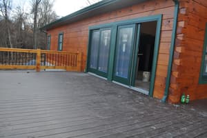 A wide plank deck running along a log wall, a sliding glass door standing open to a bare interior, with a railing at the far edge.