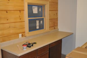 A work counter with dark cabinets and a solid countertop installed below a trimmed window set in a log wall, tools left on top.