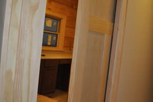 A view through an open paneled door with new trim into a tiled room holding dark cabinets and a window still masked with tape.