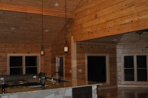 A finished great room and kitchen with a stone-faced island under granite, plank flooring, and board walls rising to a vaulted ceiling.