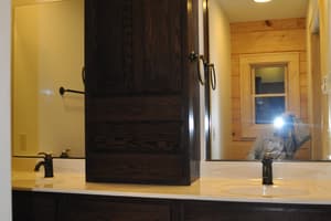 A double bathroom vanity in dark stained cabinetry with a tall center cabinet between the sinks, under recessed lights.