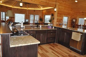 A finished kitchen with an L of dark cabinets under granite, plank flooring, and log walls opening to a dining bay under a coved tray ceiling.