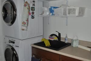 A laundry room with a stacked washer and dryer, two wall shelves, and a deep utility sink in a cabinet run with tile flooring underfoot.