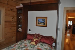 A bedroom corner where a built-in cabinet surround with narrow display shelves frames the bed, a doorway opening to the plank-floored main room.