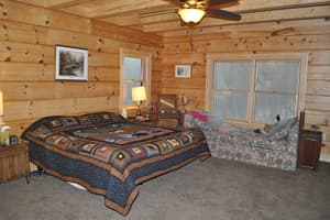 A carpeted bedroom with log walls, a board ceiling crossed by a beam, a ceiling fan, and tall windows looking out into bare woods.