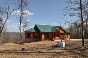 The finished log home on its ridge above a graded yard of bare earth, green metal roof over log walls, with wooded hills falling away behind.