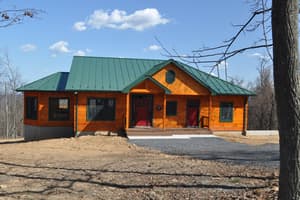 The front of the finished log home with a green metal roof, red entry doors, a covered porch, and a gravel apron across the graded yard.