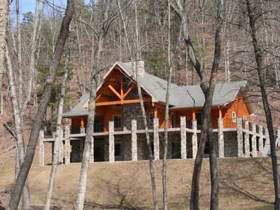 A log home in bare woods, a heavy timber truss filling the entry gable above a covered porch on stone piers