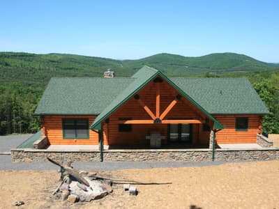 A log home square on, sunburst timber ornament centered in the gable above the entry, stone base and green roof, hills stacked behind