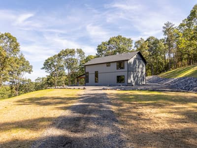 The home on its hilltop clearing seen from below, a gravel path and rock-lined bank rising toward the covered patio
