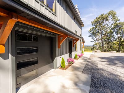 Black glass-paneled garage door under timber knee braces, with mums and young evergreens planted along the concrete walk
