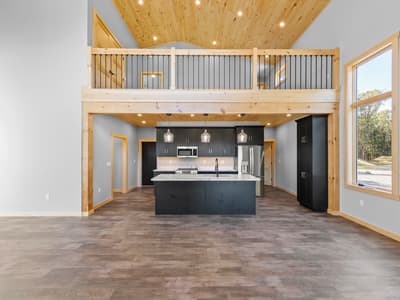 Empty great room looking toward the kitchen, with a vaulted pine ceiling, iron-spindle loft rail and a marble-look island