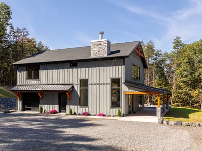 Gray board-and-batten home with a stacked-stone chimney, timber gable truss, covered entry, and flowering mums along the concrete walk