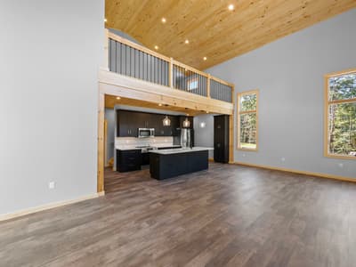 Unfurnished great room with tall windows into the woods, the kitchen and loft beyond, and gray plank flooring throughout