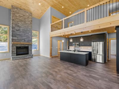 Unfurnished great room showing the stone chimney, kitchen with stainless appliances, and windows onto the gravel drive