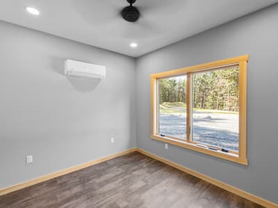 Empty bedroom with a ceiling fan, wall-mounted mini-split and a pine-trimmed window onto the gravel drive and woods