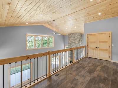 Loft with a plank pine ceiling, timber-and-iron railing, the stacked-stone chimney rising past it, and double pine doors to a bedroom
