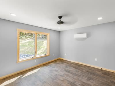 Bare bedroom with a window onto the grassy slope and rock bank, a mini-split on the wall and ceiling fan overhead