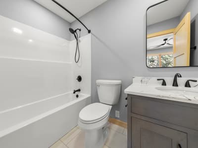 Hall bathroom with a white tub-shower, black hand shower on a rail, toilet, and dark vanity under a rounded black mirror