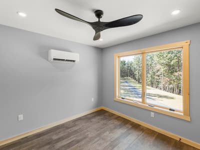 Empty bedroom with a black three-blade ceiling fan, a mini-split, and a window looking down the gravel drive through the trees
