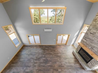 Great room seen from the loft — bare floor, a window band with wooded-hills view, mini-split, glass doors and the stone fireplace