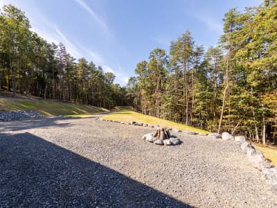 Gravel turnaround with a boulder-ringed fire pit stacked with logs, beside a mowed strip running between two tree lines