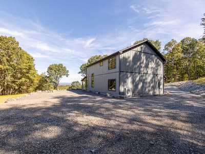 The home seen across the wide gravel parking area, a mini-split condenser against the wall and woods on three sides