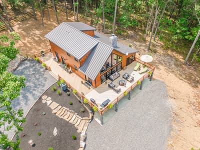 Elevated view looking down on the metal roof and wraparound deck, with cleared ground at the forest edge