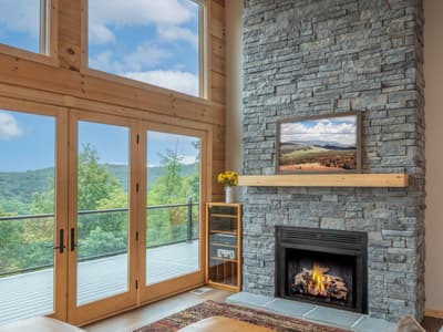Stacked-stone fireplace with a lit fire and a timber mantel, beside doors onto the deck