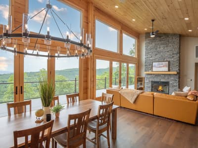 Great room with a lit stone fireplace, a tan leather sectional and a wrought-iron chandelier above the dining table
