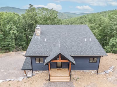 The home from in front and above, its shingle roof and stone chimney set against layered wooded ridges