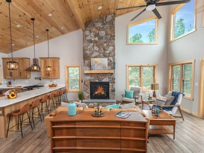 The great room seen across the kitchen island, a lit stone fireplace rising to the vaulted pine ceiling under clerestory windows