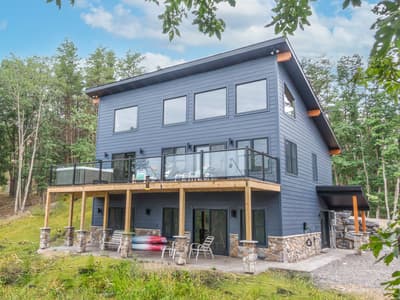 Rear elevation from ground level, three stories of siding above a deck on stone piers, forest on every side