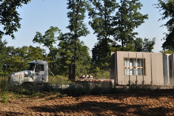 A flatbed tractor-trailer parked on the site carrying gray precast concrete wall panels stood on edge, window openings already cast into them.