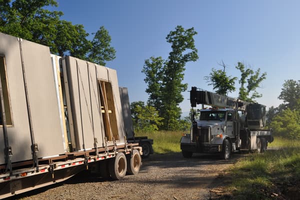 A second trailer of panels waiting behind the first, the load strapped upright, a boom truck at the end of the drive.