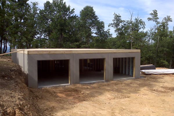 The lower level from the downhill side, three garage openings cast into the wall and the bank cut back behind.