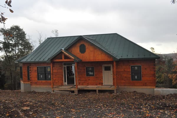 The finished exterior in late fall, stained log walls under a green metal roof, leaves down and the ground bare.
